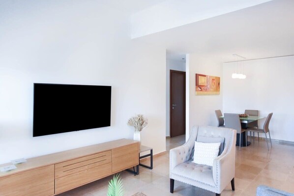 A modern living room with a flat-screen TV mounted on the wall, a wooden console table, a light-colored armchair, and a dining area with a table and chairs.