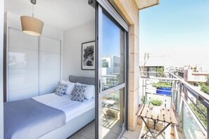 Un dormitorio moderno con cama, lámpara colgante, cuadro enmarcado y balcón con mesa y barandilla.