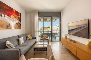 Una sala de estar moderna con un sofá gris, una mesa de centro y un aparador de madera. Hay un cuadro grande en la pared y una pantalla de televisión mostrando un paisaje.