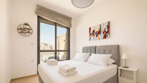 Habitación de hotel con cama, mesita de noche, lámpara, un cuadro y ventana con vistas a edificios.