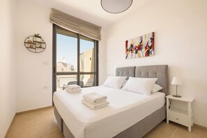 Habitación de hotel con cama, mesita de noche, lámpara, un cuadro y ventana con vistas a edificios.