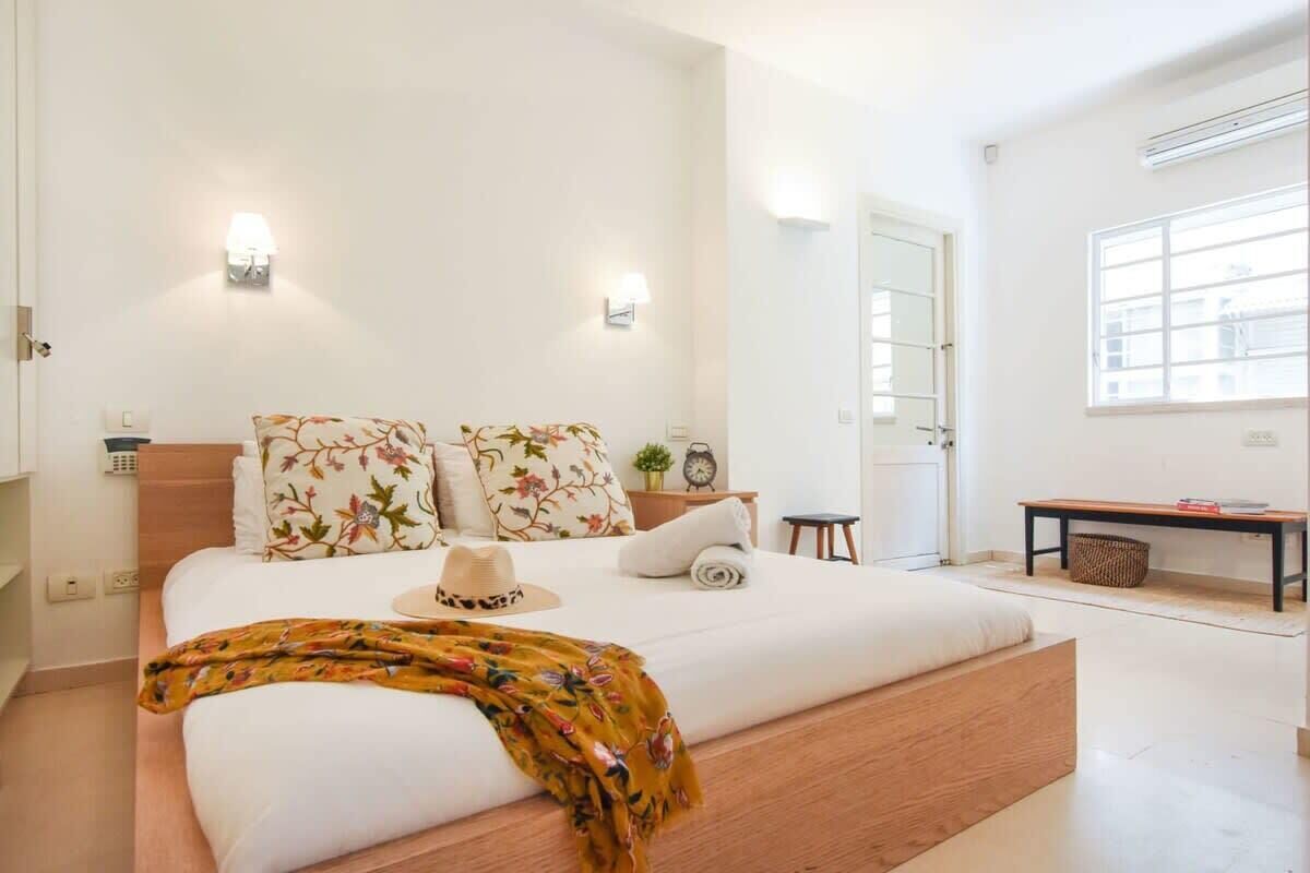 A modern bedroom with a large bed, floral-patterned pillows, a wooden headboard, bedside tables with lamps, and a window providing natural light.