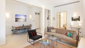 A modern living room with a white brick wall, a sofa, a coffee table, and a dining area with chairs and a table. - Spacious 3BR at Mapu st by HolyGuest (Tel Aviv)