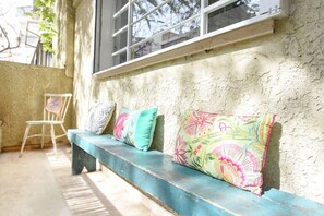 Un banc agrémenté de coussins aux couleurs vives, adossé à un mur à la texture intéressante, une chaise et une fenêtre avec des stores.