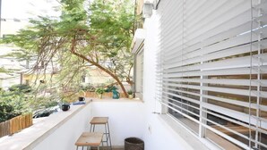 A narrow balcony with a white railing, a small wooden table, a wicker basket, and a window with white blinds. - Spacious 3BR at Mapu st by HolyGuest (Tel Aviv)