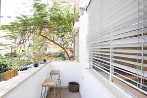 A narrow balcony with a white railing, a small wooden table, a wicker basket, and a window with white blinds.