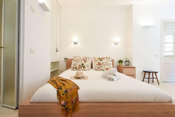 A neatly made bed with floral bedding, a bedside table with a clock, and a small stool. - Spacious 3BR at Mapu st by HolyGuest (Tel Aviv)