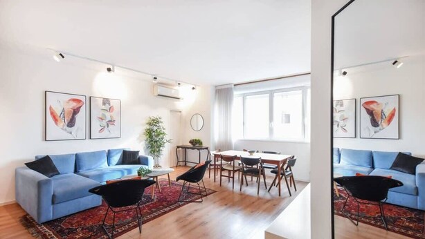 A modern living room with a blue sofa, a dining table, and a large window. - Charming 2BR in Mapu  by HolyGuest (Tel Aviv)
