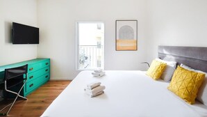 A hotel room with a bed, a television mounted on the wall, a window with a view of buildings, and a door leading to an outdoor balcony. - Charming 2BR in Mapu  by HolyGuest (Tel Aviv)