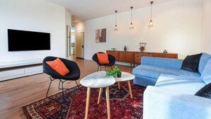 A modern living room with a blue sofa, black chairs, a coffee table, and a flat-screen TV mounted on the wall. - Charming 2BR in Mapu  by HolyGuest (Tel Aviv)