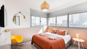 A modern hotel room with a large bed, a yellow chair, a small table, and a large window offering a city view. - Modern 3-BR near Beach Club by HolyGuest (Tel Aviv)