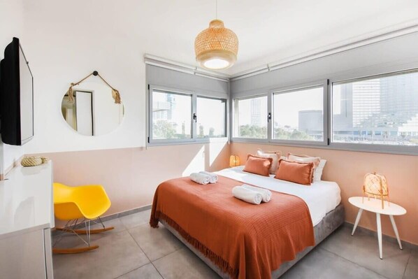 Habitación de hotel moderna con una cama grande, una silla amarilla, una mesita pequeña y un ventanal con vistas a la ciudad.