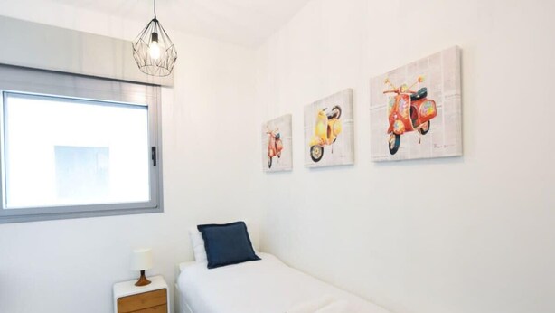 A bedroom with a bed, a nightstand, a lamp, and three wall-mounted paintings of scooters. - Modern 3-BR near Beach Club by HolyGuest (Tel Aviv)