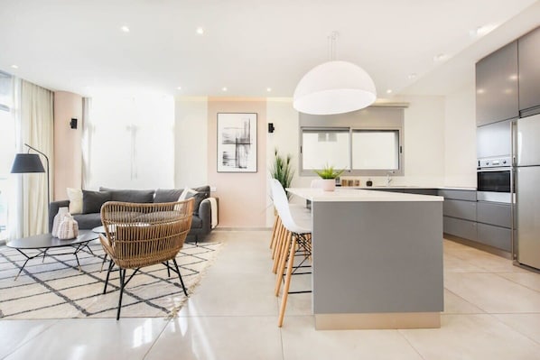 A modern kitchen with a central island, a dining area with chairs, a sofa, and a lamp.