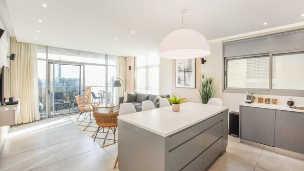 A modern apartment with a central island, a dining area with wicker chairs, a large window, and a balcony with a view. - Modern 3-BR near Beach Club by HolyGuest (Tel Aviv)