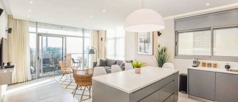 A modern apartment with a central island, a dining area with wicker chairs, a large window, and a balcony with a view.