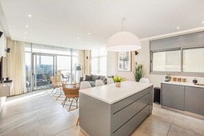 A modern apartment with a central island, a dining area with wicker chairs, a large window, and a balcony with a view.