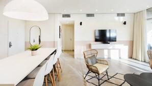 A modern living room with a white sofa, a dining table with chairs, a large mirror, and a flat-screen TV on a white console. - Modern 3-BR near Beach Club by HolyGuest (Tel Aviv)