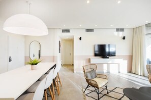 A modern living room with a white sofa, a dining table with chairs, a large mirror, and a flat-screen TV on a white console.