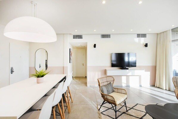 A modern living room with a white sofa, a dining table with chairs, a large mirror, and a flat-screen TV on a white console.