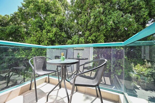 A glass-enclosed balcony with two tables and four chairs, surrounded by lush green trees. - Bright 2BR Apartment with Balcony in Dizengoff by HolyGuest (Tel Aviv-Yafo)