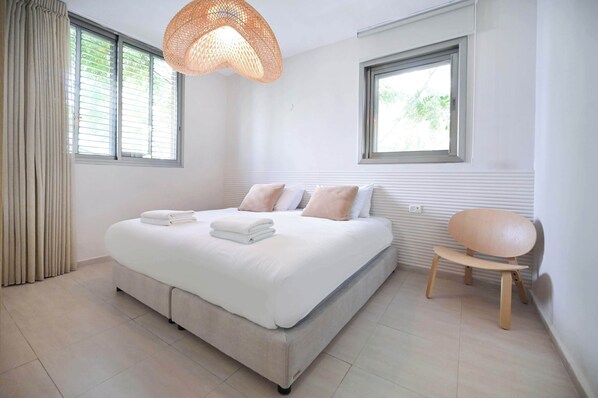 A modern bedroom with a large bed, two windows, a wooden chair, and a woven light fixture.