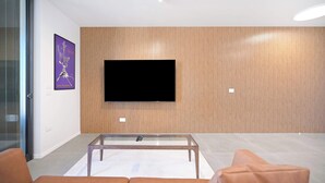 A modern living room with a flat-screen TV mounted on a wooden wall, a glass coffee table, and orange leather sofas.