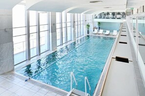 En indendørs swimmingpool med glasvægge, flisegulv og et loungeområde med liggestole.