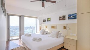 Une chambre moderne avec un grand lit, un ventilateur de plafond, une table de chevet et une vue sur la ville grâce à de grandes fenêtres.
