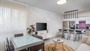 A modern living room with a glass dining table, a sofa, a TV, and a kitchen area with bar stools. - Spacious 3BR on Frishman Beach by HolyGuest (Tel Aviv)