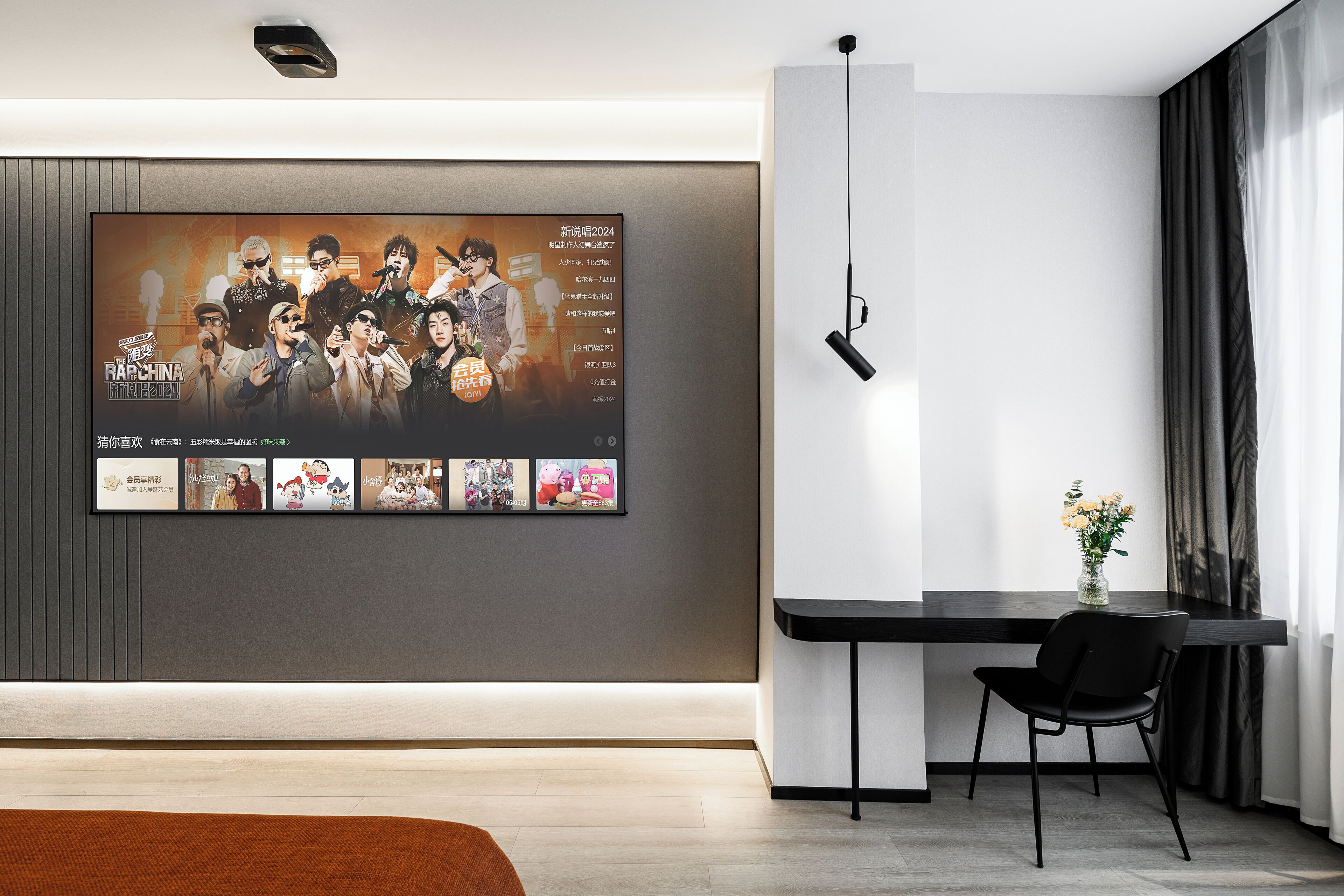 A modern hotel room with a large flat-screen TV mounted on the wall displaying a music band, a small table with a vase of flowers, and a black chair.