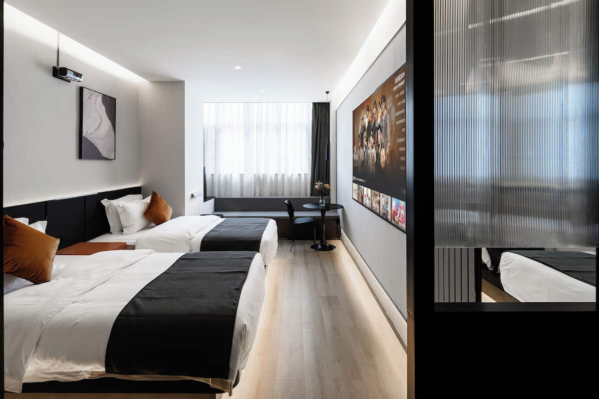 A hotel room with two beds, a small table, and a large window with sheer curtains.