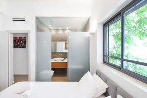 A modern bedroom with a large window, a bed with white bedding, and a view of a bathroom with a sink and mirror. - Magic Lily in Frug by HolyGuest (Tel Aviv)