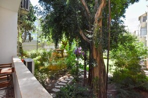 A balcony with a stone pathway, lush greenery, and a large tree.