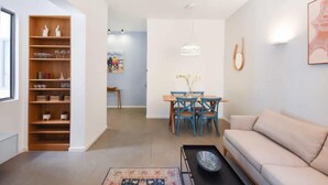 Un salon moderne avec un canapé beige, une table basse, un coin repas comprenant une table en bois et des chaises bleues, ainsi qu’une bibliothèque agrémentée d’objets décoratifs.