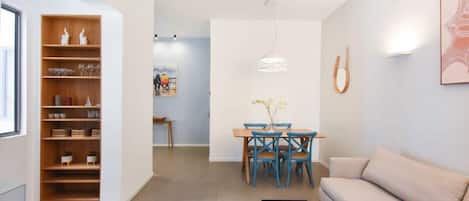 Un salon moderne avec un canapé beige, une table basse, un coin repas comprenant une table en bois et des chaises bleues, ainsi qu’une bibliothèque agrémentée d’objets décoratifs.