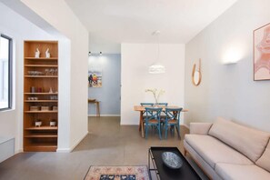 A modern living room with a beige sofa, a coffee table, a dining area with a wooden table and blue chairs, and a bookshelf with decorative items. - Magic Lily in Frug by HolyGuest (Tel Aviv)