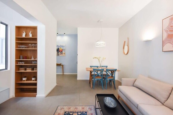 Una sala de estar moderna con un sofá color beige, una mesa de centro, un comedor con una mesa de madera y sillas azules, y una estantería con objetos decorativos.