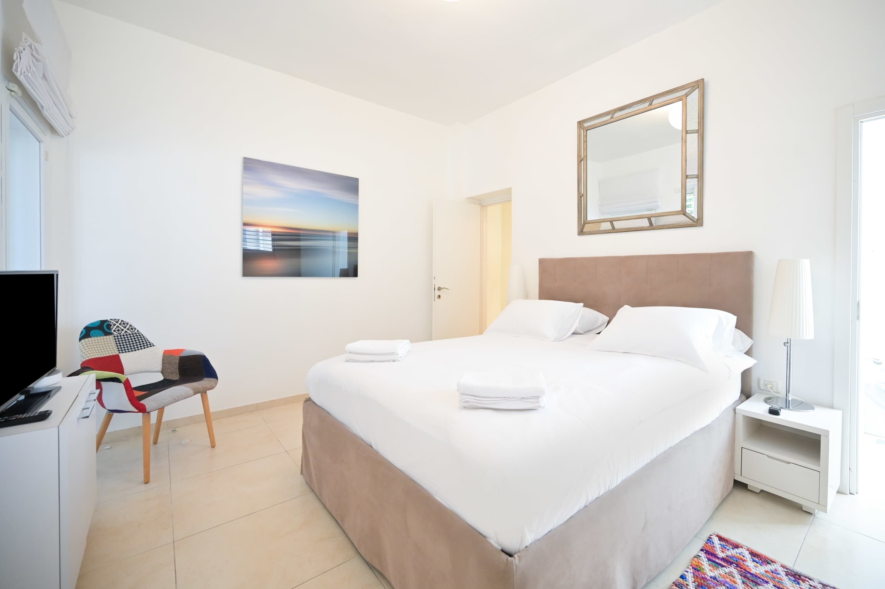 Ein Hotelzimmer mit Bett, Schreibtisch mit Computer, Stuhl, Fernseher, Spiegel und einem Gemälde an der Wand.