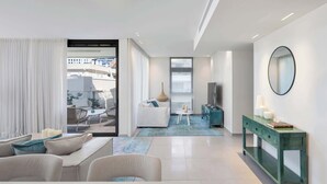 Een moderne woonkamer met een bank, een salontafel en een televisie. Er is een balkon met witte gordijnen en uitzicht op gebouwen.