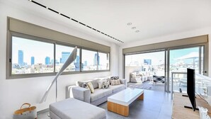 A modern living room with a large window, a sofa, a coffee table, and a TV. - Luxury 3BR Beachfront Penthouse by HolyGuest (Tel Aviv)
