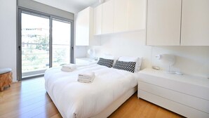 A modern bedroom with a large bed, white cabinets, a bedside table with a lamp, and a view through a sliding glass door. - Luxury 3BR Beachfront Penthouse by HolyGuest (Tel Aviv)