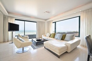 A modern living room with a large sofa, armchairs, a flat-screen TV, and large windows offering a view of the beach. - Luxurious Beach Front 1BR by HolyGuest (Tel Aviv)