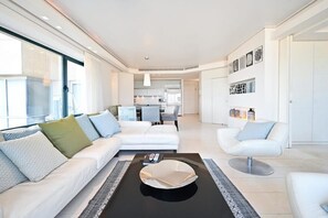 A modern living room with a sofa, armchair, and coffee table. A kitchen is visible in the background.