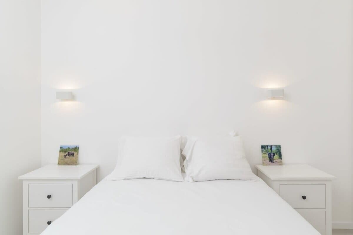 Un dormitorio minimalista con una cama individual, ropa de cama blanca, dos mesitas de noche con luces fijadas a la pared y pequeños cuadros enmarcados sobre las mesitas.