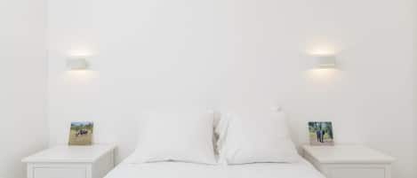 Un dormitorio minimalista con una cama individual, ropa de cama blanca, dos mesitas de noche con luces fijadas a la pared y pequeños cuadros enmarcados sobre las mesitas.