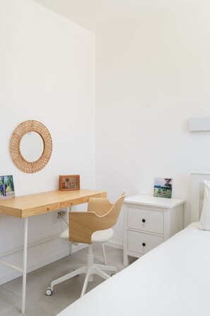 Un dormitorio minimalista con un escritorio de madera, un tapiz de pared tejido, un cuadro enmarcado, una mesita de noche blanca y una cama.