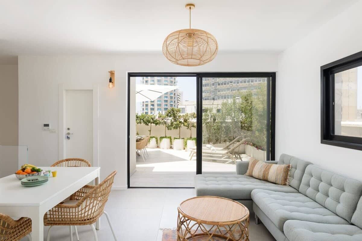 A modern living room with a sofa, dining table, and chairs. There is a large window with a view of buildings and greenery.