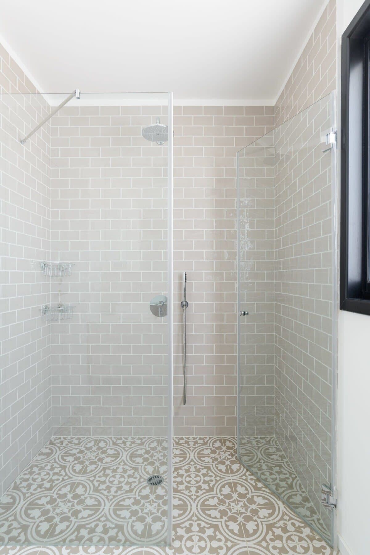 A shower with clear glass doors, white tiled walls, and patterned floor tiles.