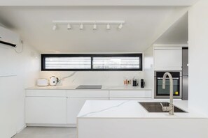 A modern kitchen with white cabinetry, a marble countertop, a built-in oven, and a window with blinds.